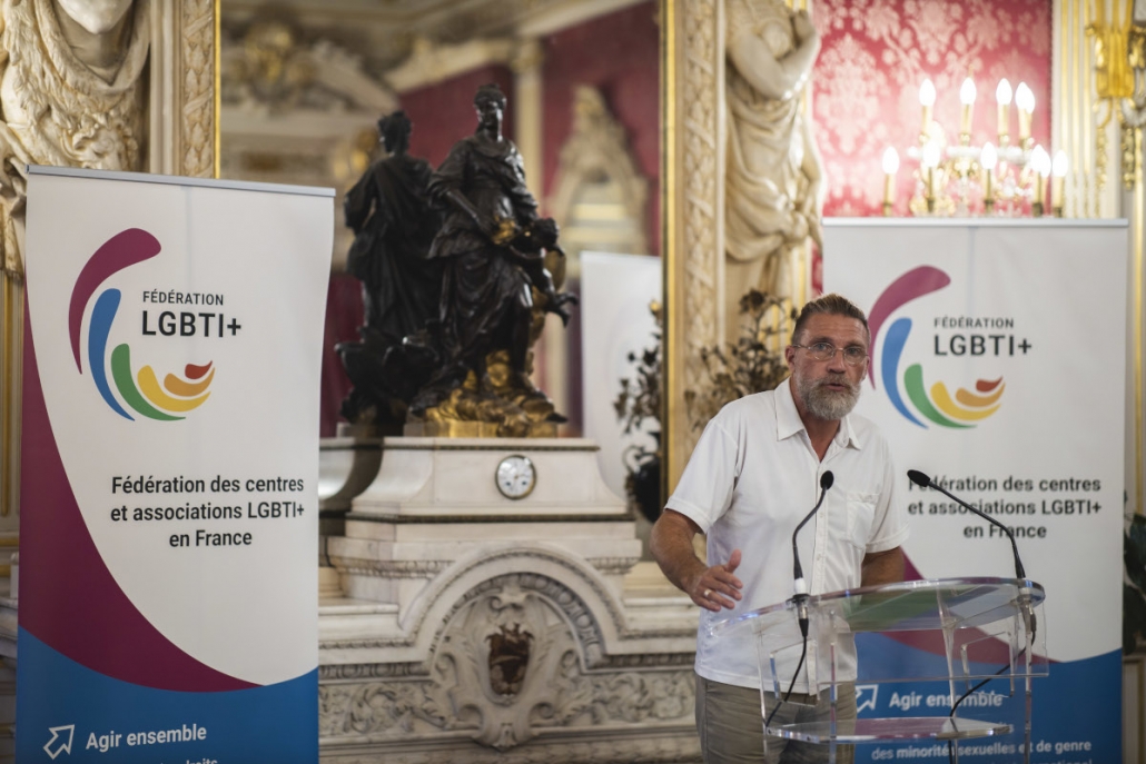 Photographie de M. Jean-Christophe TESTU, président de la Fédération LGBTI+, devant un pupitre dans une salle richement décorée de la Mairie centrale de Lyon, flanqué de deux kakémonos de la Fédération LGBTI+